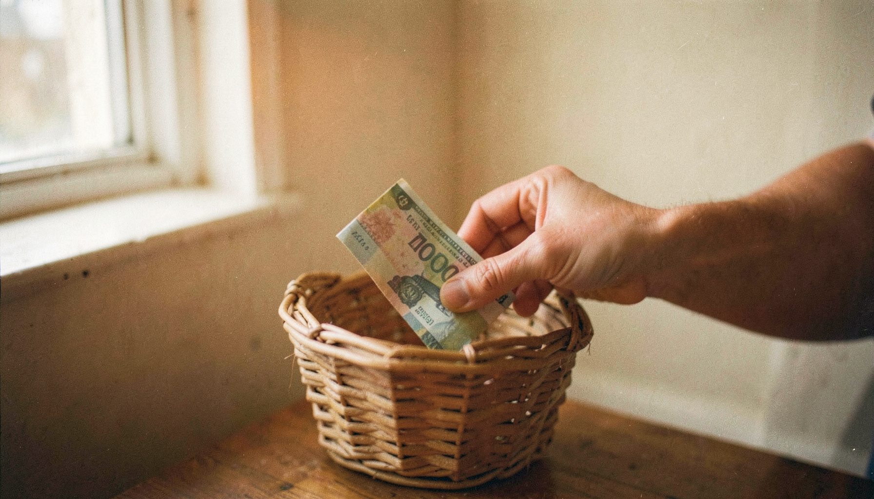 Warm-toned analog film photograph of hands gently placing a folded banknote into a simple woven basket, soft natural light from a nearby window, muted earth tones and cream walls, visible grain.