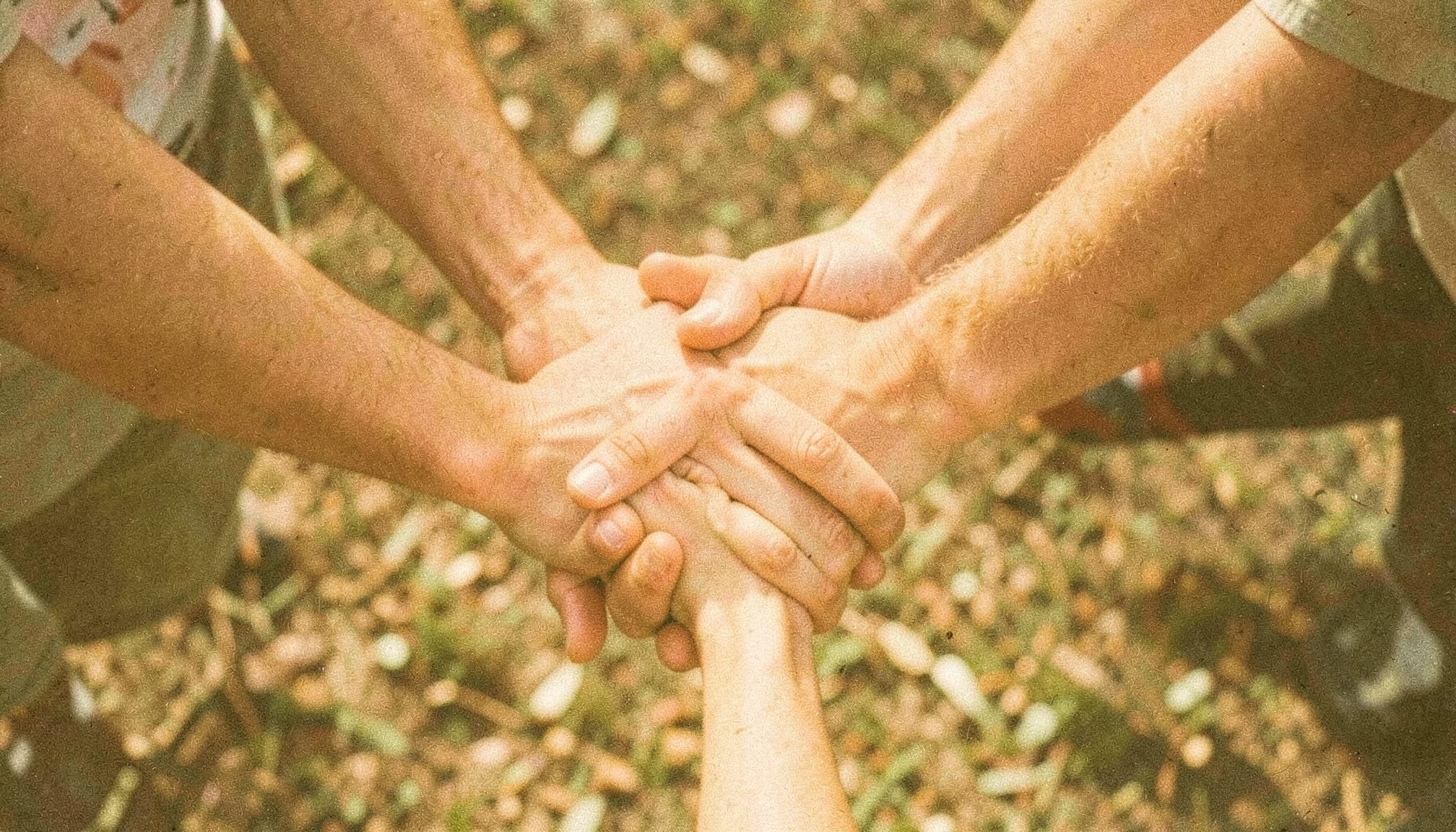 A warm, grainy film photograph of hands joined together in a circle, symbolizing community service and fellowship, with soft golden and olive tones against an earthy background.